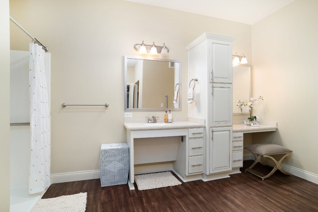 CelebrationVillageLg-50 - Celebration Village Forsyth A photo of the bathroom with a sink, two mirrors, a vanity table and chair, and a walk-in shower at Celebration Village Forsyth, a senior living community near Cumming, GA