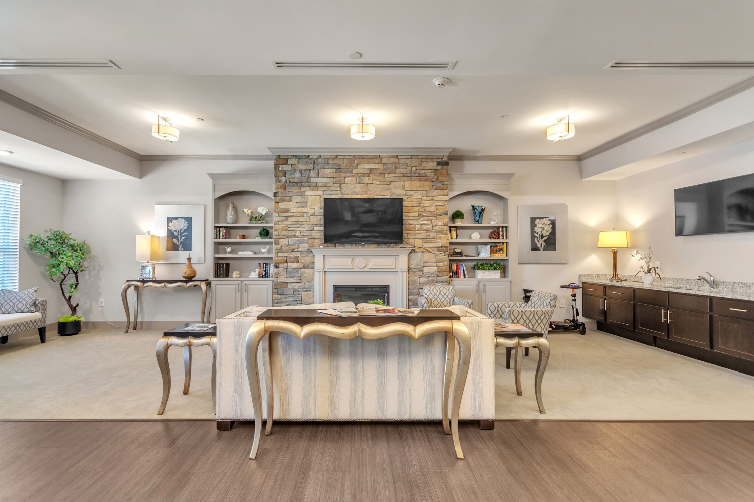 A room with two TVs, fireplace, couch, sofa chairs, multiple desks, a sink, and brown cabinets  at Celebration Village Forsyth, a senior living community near John's Creek, GA.