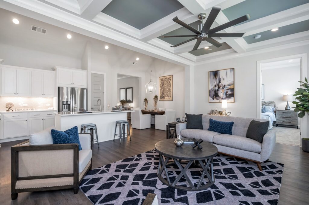 (13) Cottage Main Living Large - Celebration Village Forsyth A photo of the large Cottage Main living room with a sofa, coffee table, fan, and a connected kitchen with a kitchen island and bar stools at Celebration Village Forsyth, a senior living community near Alpharetta, GA.